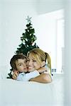 Knaben und Mutter auf Sofa umarmen einander, Weihnachtsbaum im Hintergrund