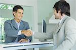 Young man shaking hands with mature businessman across desk, smiling