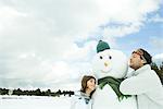 Brother and sister leaning against snowman, both smiling, brother's eyes closed