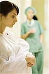 Young woman holding towel in hospital corridor, looking down, medical worker in background