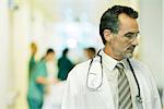 Male doctor looking away, head and shoulders, hospital corridor in background