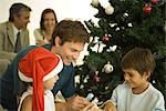 Father and two children sitting by Christmas tree, opening presents together, daughter wearing Santa hat