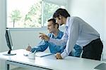 Businessman sitting at desk facing monitor while colleague stands next to him, speaking and pointing to screen