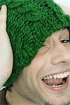 Jeune homme portant chapeau, un oeil couvert, riant de caméra en tricot