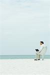 Businessman sitting in chair on beach, using laptop computer, looking away