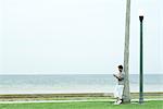 Man leaning against tree trunk, looking down at cell phone, full length