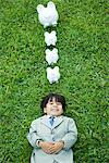 Little boy in full suit lying on grass, line of piggy banks above head