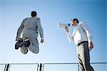 Businessman standing beside colleague, shouting into megaphone, colleague jumping in air, low angle view