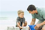 Father and son playing with sand bucket at the beach, smiling at each other