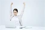 Young businesswoman sitting at desk, smiling at laptop and raising both arms in the air