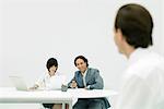 Professionals sitting at desk, woman reading document, man looking at person in foreground