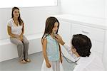 Pediatrician holding stethoscope to girl's chest, mother sitting nearby