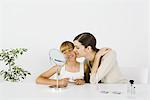 Young woman and preteen sister sitting in front of mirror, older sister kissing younger sister's cheek