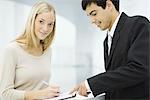 Businessman holding out clipboard, woman writing on document and smiling at camera