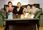 Three young people and one child sitting on green couch together, laughing, blurred rear view of tv in foreground