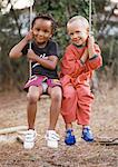 Little boy and girl sitting on swing together, each holding one side of swing rope, smiling at camera, full length.