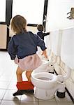 Little girl next to children's toilet with panties around ankles, rear view, other toilets and child in background.