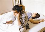 Man sitting up in bed, pointing remote control, woman lying behind him.