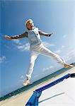 Mature woman jumping on trampoline at the beach, arms out, legs spread