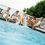 Young people sitting around swimming pool, splashing feet in water