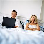 Couple in bed, man using laptop computer, woman with arms crossed