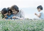 Man lying on grass with boy and girl looking at cellphone, second boy sitting apart