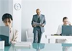 Male executive standing in office reading newspaper, female colleagues working on laptop computers