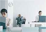 Two women working at laptops in office, businessman sitting in chair reading newspaper in background