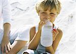 Boy sitting on sand next to mother, drinking water