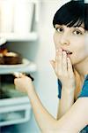 Young woman taking slice of cake from refrigerator, looking at camera, covering mouth