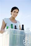 Woman carrying recycling bin filled with glass bottles, smiling at camera