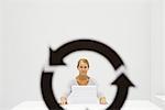 Professional woman sitting behind stack of paper, looking at camera through recycling symbol