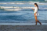 Junge Frau mit Handy, beim Spaziergang am Strand, Seitenansicht