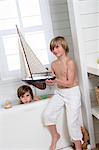 2 boys in bathroom, one having a bath, the other holding a model boat