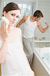 Couple in a bathroom, woman cleansing face with cotton wool, man flexing muscles in background