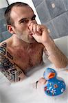 Tattooed man having a bath, sucking his thumb, holding a plastic duck