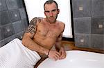 Tattooed man, barechested, lying on tub edge, looking at the camera