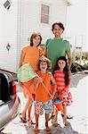 Parents and two children in front of a house, holding beach accessories, outdoors