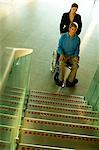 Male patient sitting in a wheelchair and a mid adult woman standing beside him near a staircase