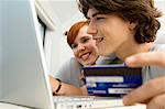 Close-up of a teenage boy holding a credit card and using a laptop with a young woman sitting beside him