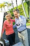 Man talking on a mobile phone beside his wife loading a luggage into car