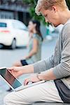 Jeune homme avec une femme en arrière-plan sur une rue à l'aide d'un ordinateur portable