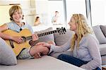 Teenage boy playing a guitar with his mother sitting near him and smiling