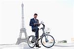 Homme d'affaires, tenir un journal et un mobile sur un vélo avec la tour Eiffel en arrière-plan, Paris, Ile-de-France, France