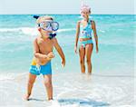 Happy young boy with snorkeling equipment on sandy tropical beach, his sister background.