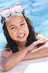 A cute happy young interracial African American girl child relaxing on the side of a swimming pool smiling & wearing pink goggles