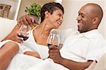 A happy African American man and woman couple in their thirties sitting at home together laughing and drinking glasses of red wine.