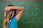 Little schoolgirl thinking while scratching the back of her head in front of a blackboard