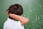 Schoolboy thinking while scratching the back of his head in front of a chalkboard