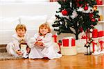 Two twins girl sitting with presents near Christmas tree